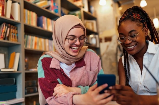 A Muslim student in a pink hijab shows her phone to another student, both smiling.